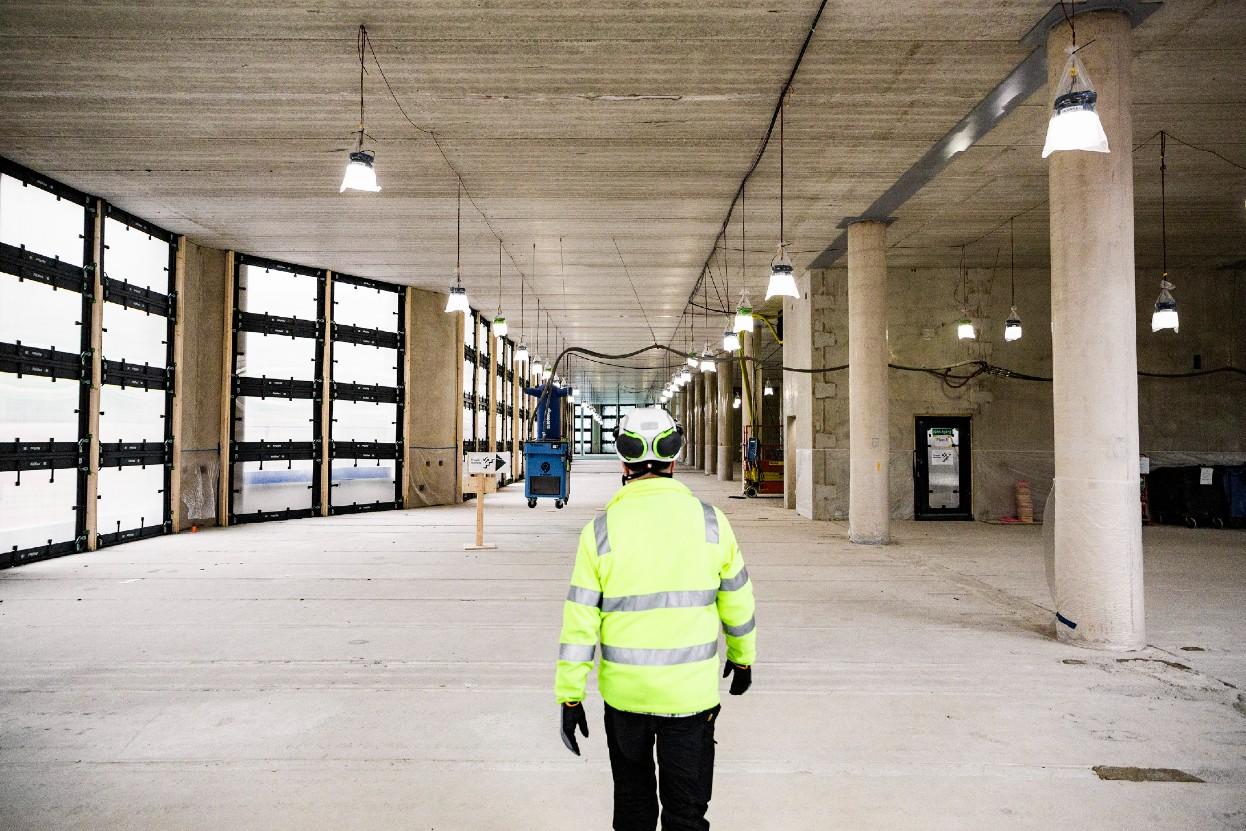 Byggarbetare i gul varseljacka och hjälm sedd bakifrån på en byggarbetsplats inomhus, med rader av arbetsbelysning och stora fönsterpartier.